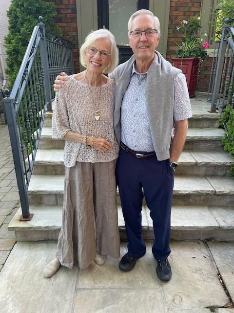 Jan and Tom, smiling in front of some steps outside. Jan is wearing a tan coloured shirt and skirt and Tom is wearing a light blue shirt, grey sweater around his shoulders, and blue pants..
